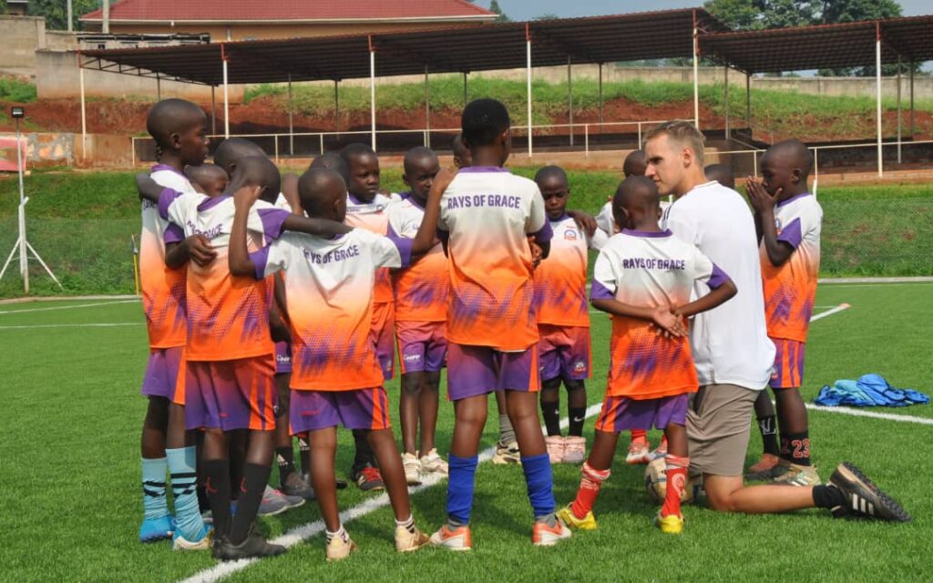 Ik zit op één knie als onderdeel van een cirkel van jonge spelers bij Rays of Grace Academy en geef uitleg tijdens een voetbaltraining.